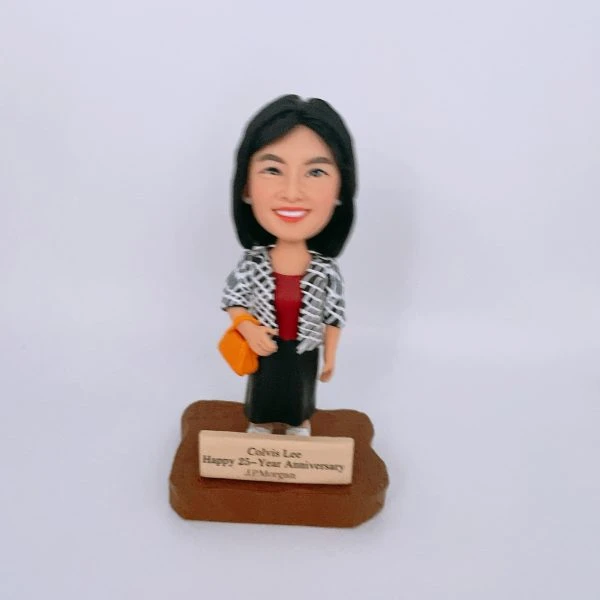 一款女性人像公仔站在木質感不規則底座上，前方金色銘牌刻有祝賀25週年與公司名稱字樣；公仔黑色短髮、微笑表情，身穿酒紅色上衣、黑色長裙與黑白格紋外套，右手挽橙黃色手袋，腳穿淺色鞋。背景純白，整體呈公司獎座風格的人像公仔。
