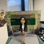 黑長髮戴眼鏡的女性人像公仔，身穿黑色西裝外套與白色上衣，搭配黑白橫條紋窄裙與黑色高跟鞋；雙手於身前交握、端正站立並微笑。公仔置於灰色粗紋質感底座，兩側有綠色點綴，背板為綠色遮陽棚與玻璃門的街景店面，周圍有綠植與牆面細節。