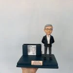 同事退休主題的人像公仔：銀灰短髮男性穿深藍西裝外套、白襯衫與灰色長褲，雙手插袋微笑站立；深藍色底座前方有金色名牌刻上姓名、職稱與「Happy Retirement 2023」。底座左側結合雙槽卡片座，可放名片或小卡，內置示範名片。整體呈現辦公室桌上型退休禮物的人像公仔。