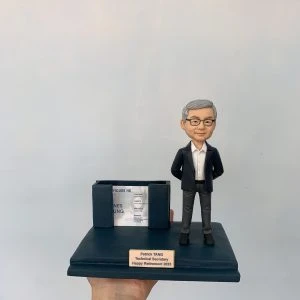 同事退休主題的人像公仔：銀灰短髮男性穿深藍西裝外套、白襯衫與灰色長褲，雙手插袋微笑站立；深藍色底座前方有金色名牌刻上姓名、職稱與「Happy Retirement 2023」。底座左側結合雙槽卡片座，可放名片或小卡，內置示範名片。整體呈現辦公室桌上型退休禮物的人像公仔。