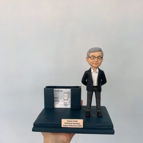 同事退休主題的人像公仔：銀灰短髮男性穿深藍西裝外套、白襯衫與灰色長褲，雙手插袋微笑站立；深藍色底座前方有金色名牌刻上姓名、職稱與「Happy Retirement 2023」。底座左側結合雙槽卡片座，可放名片或小卡，內置示範名片。整體呈現辦公室桌上型退休禮物的人像公仔。