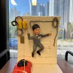 一座情景式人像公仔：男性角色穿灰色連身工作服，胸前貼有「JPL」字樣貼章，雙臂外伸、雙腳彎曲，呈失重漂浮姿態，固定在白色鉚釘結構艙壁前。艙壁左上方掛有黃色裝置與黑色電線，旁邊繞著灰色管線；右側亦有環狀纜線。地面左前方放置以白色綁帶固定的紅黑色軟包裝備。整體置於木桌上，背景是窗外的高樓城市景色與藍天。