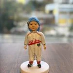 放在木桌上的小朋友主題人像公仔：bb頭戴淺藍棒球帽反戴，身穿白底灰細條的棒球連身服，胸前有紅字「ALL-Star」與小盾牌章；腳穿紅黃條紋襪與黑色卡通圖案鞋，站在白色圓形底座上；窗外背景為散景的高樓與街景。