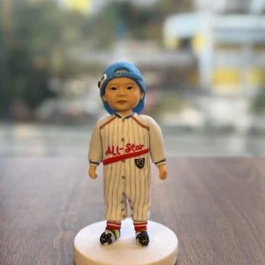 放在木桌上的小朋友主題人像公仔：bb頭戴淺藍棒球帽反戴，身穿白底灰細條的棒球連身服，胸前有紅字「ALL-Star」與小盾牌章；腳穿紅黃條紋襪與黑色卡通圖案鞋，站在白色圓形底座上；窗外背景為散景的高樓與街景。