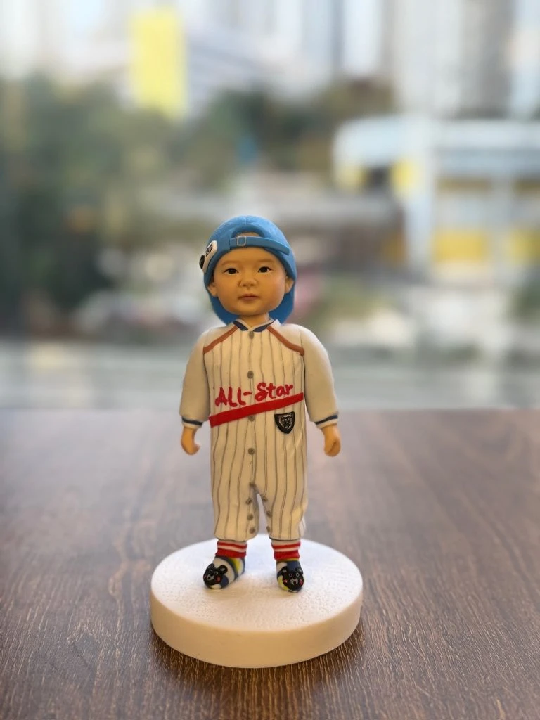 放在木桌上的小朋友主題人像公仔：bb頭戴淺藍棒球帽反戴，身穿白底灰細條的棒球連身服，胸前有紅字「ALL-Star」與小盾牌章；腳穿紅黃條紋襪與黑色卡通圖案鞋，站在白色圓形底座上；窗外背景為散景的高樓與街景。