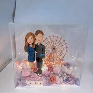 透明展示盒中的情侶人像公仔場景：左側長髮角色穿深藍色連身裙與白鞋，右側短髮角色穿深灰色西裝與黑鞋，兩人相互依偎站立。背景是一座金銅色摩天輪模型，周圍環繞暖白色小燈串。底部鋪滿乾燥花與仿真花材，色系涵蓋粉、玫瑰紅、奶白與淡紫；前方名牌寫有「Kit & HY」。整體呈現夢幻溫暖的約會氛圍。