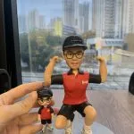 一組人像公仔擺設放在木桌上：主體公仔戴黑色鴨舌帽（上有「Superme」字樣）、黑框眼鏡，穿紅黑配色短袖上衣左胸印有「KMB」字樣、黑色短褲與白色運動鞋；姿勢為單膝下蹲並雙臂彎曲展示肌肉。旁邊手指捏著一個迷你版吊飾公仔，服飾與帽款同款。兩者共同置於灰色圓形底座，背景是玻璃窗外的高樓城市景觀。