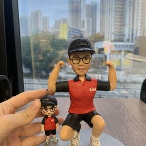 一組人像公仔擺設放在木桌上：主體公仔戴黑色鴨舌帽（上有「Superme」字樣）、黑框眼鏡，穿紅黑配色短袖上衣左胸印有「KMB」字樣、黑色短褲與白色運動鞋；姿勢為單膝下蹲並雙臂彎曲展示肌肉。旁邊手指捏著一個迷你版吊飾公仔，服飾與帽款同款。兩者共同置於灰色圓形底座，背景是玻璃窗外的高樓城市景觀。