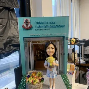 一款老師主題的人像公仔，置於校門口場景底座上。公仔留中長黑髮，穿白色上衣與藍花圖案裙，手抱金色獎盃（杯身有中文「最佳老師」字樣）；腳邊擺放編織花籃，內有多色立體花朵。背景板為幼稚園入口與校徽，上方英文與中文寫有幼稚園名稱及年份。地台為灰色地面，兩側以綠色草地條紋點綴。整體陳列在室內桌面，周邊可見辦公與教具物品。