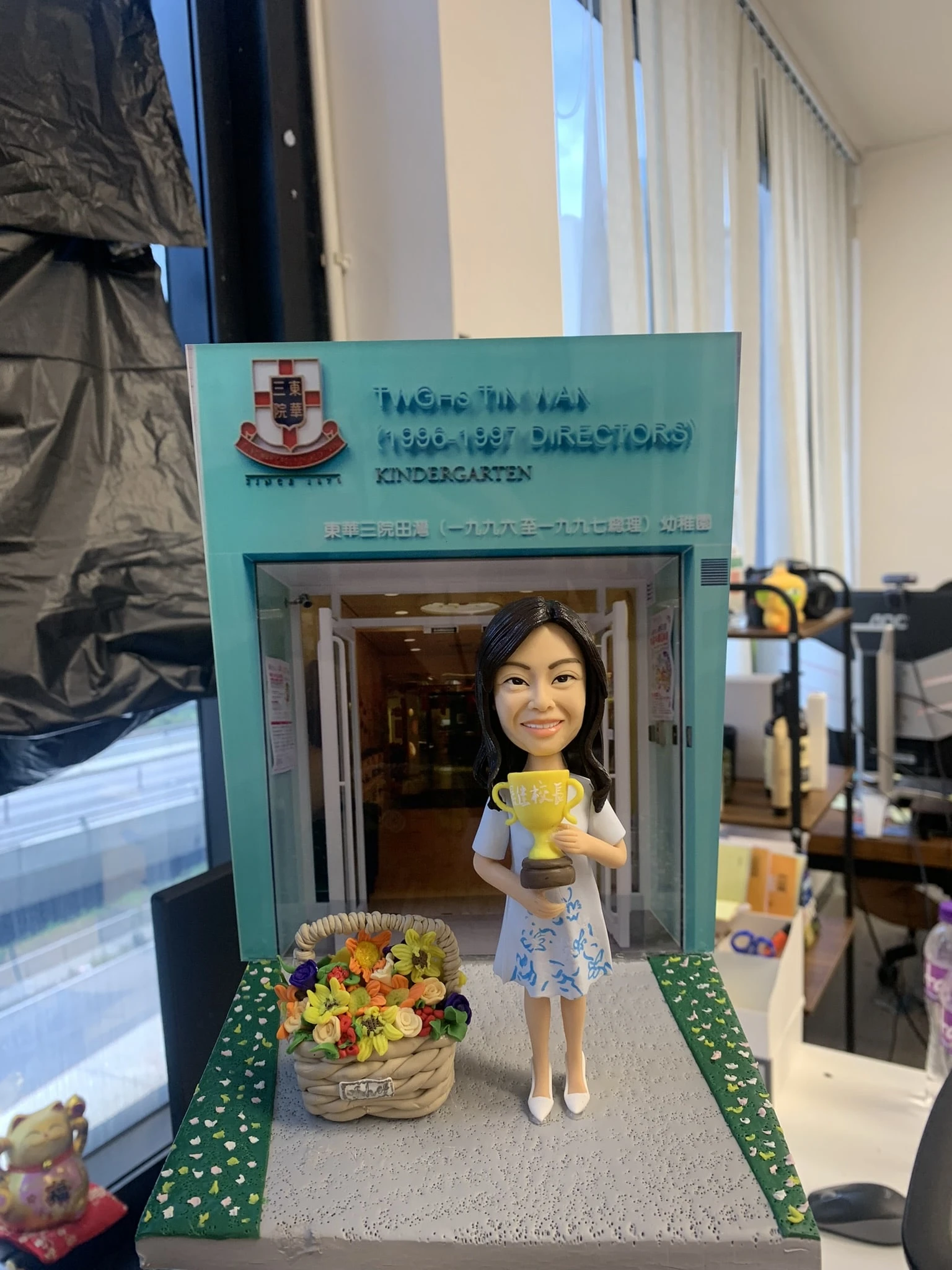 老師主題人像公仔，手抱金色獎盃與花籃，立於幼稚園門口場景