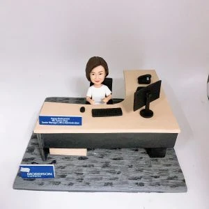 辦公室主題人像公仔坐在L形辦公桌後方，身穿白色上衣，面前有鍵盤與滑鼠，右側擺放雙螢幕顯示器與桌上電話。桌沿藍色名牌寫有「Happy Retirement… Senior Manager, HR & Administration」祝福與職稱。底座前方有「MORRISON EXPRESS」藍白標誌牌。整體呈現人資與行政管理場景的人像公仔擺設。