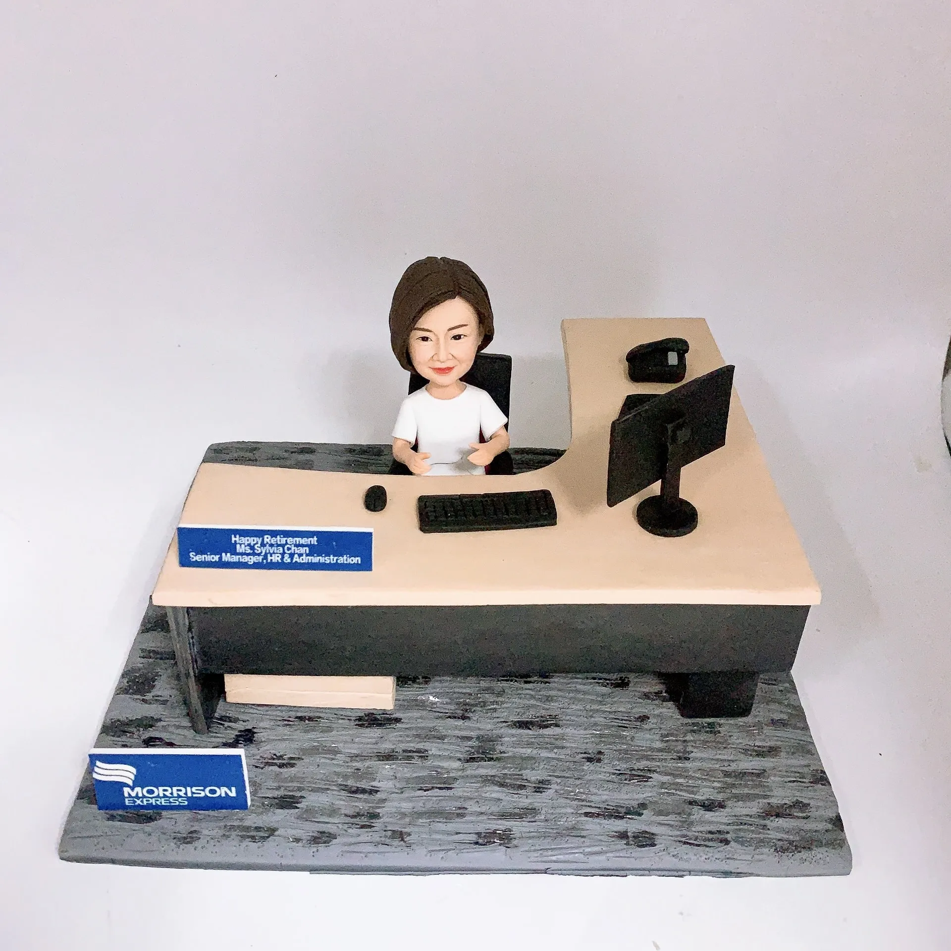 辦公室主題人像公仔坐在L形辦公桌後方，身穿白色上衣，面前有鍵盤與滑鼠，右側擺放雙螢幕顯示器與桌上電話。桌沿藍色名牌寫有「Happy Retirement… Senior Manager, HR & Administration」祝福與職稱。底座前方有「MORRISON EXPRESS」藍白標誌牌。整體呈現人資與行政管理場景的人像公仔擺設。