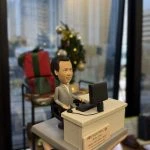 【同事退休禮物】港鐵主題的人像公仔：男同事身穿淺灰西裝與白襯衫，坐在黑色辦公椅上，雙手置於鍵盤前使用電腦；桌面配置螢幕、鍵盤與有線耳麥。米灰色辦公桌前貼有「mtr propertym」與退休祝賀名牌，底座為階梯式平台。背景可見節日裝飾與禮物盒，呈現辦公室內為港鐵同事準備的人像公仔退休禮物。