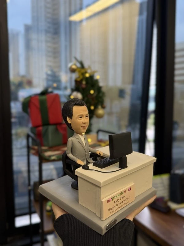 【同事退休禮物】港鐵主題的人像公仔：男同事身穿淺灰西裝與白襯衫，坐在黑色辦公椅上，雙手置於鍵盤前使用電腦；桌面配置螢幕、鍵盤與有線耳麥。米灰色辦公桌前貼有「mtr propertym」與退休祝賀名牌，底座為階梯式平台。背景可見節日裝飾與禮物盒，呈現辦公室內為港鐵同事準備的人像公仔退休禮物。