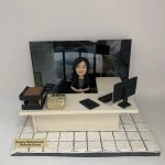 辦公室主題的人像公仔場景：黑長髮女性公仔坐在米色辦公桌後方，雙手自然放於桌上、面帶微笑。桌面擺有電腦螢幕、鍵盤、滑鼠、文件盤、計算機與文具盒；右上方有淺色側桌。地台為白色石紋地磚，前方金色名牌寫有「Happy Retirement」與名字字樣。背板為辦公室高樓窗景與工位環境照片，整體呈現OFFICE氛圍的退休致意擺設。