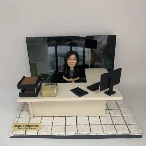 辦公室主題的人像公仔場景:黑長髮女性公仔坐在米色辦公桌後方,雙手自然放於桌上、面帶微笑。桌面擺有電腦螢幕、鍵盤、滑鼠、文件盤、計算機與文具盒;右上方有淺色側桌。地台為白色石紋地磚,前方金色名牌寫有「Happy Retirement」與名字字樣。背板為辦公室高樓窗景與工位環境照片,整體呈現OFFICE氛圍的退休致意擺設。