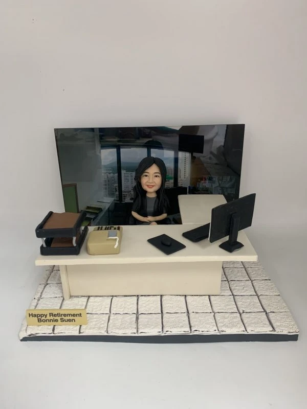 辦公室主題的人像公仔場景：黑長髮女性公仔坐在米色辦公桌後方，雙手自然放於桌上、面帶微笑。桌面擺有電腦螢幕、鍵盤、滑鼠、文件盤、計算機與文具盒；右上方有淺色側桌。地台為白色石紋地磚，前方金色名牌寫有「Happy Retirement」與名字字樣。背板為辦公室高樓窗景與工位環境照片，整體呈現OFFICE氛圍的退休致意擺設。