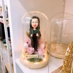 一位女性人像公仔置於透明玻璃罩內，腳下為木質底座與環形暖光燈。公仔穿著黑色長袍搭配深綠領緣，雙手比出手指愛心手勢，面帶微笑。玻璃罩內以粉色與淡紫玫瑰、苔蘚與乾燥花裝飾，前方小牌寫有「Best Secretary Yannie」與愛心圖案。背景為陳列層架與其他擺件，整體呈現溫馨致謝與頒獎紀念風格的人像公仔擺設。