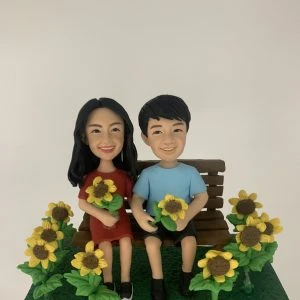 一組情侶人像公仔坐在木質長椅上，置於深綠草地質感底座中央；左側角色穿紅色連衣裙、手抱一束太陽花，右側角色穿淺藍上衣與黑色短褲、手持一朵太陽花。草地兩側與前方環繞多株盛開的太陽花，花心棕色、花瓣亮黃，整體呈現花田氛圍。