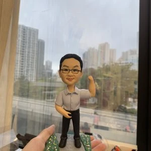 老闆主題的人像公仔立於灰色步道與綠地花卉點綴的底座上，由手掌托起展示。公仔為短髮、配戴眼鏡，穿淺灰短袖襯衫、黑色長褲與黑色皮鞋；右臂上舉作打氣姿勢，左手自然放置。背景為臨窗的辦公環境，可見城市高樓與道路景觀，前景擺有文具收納盒。