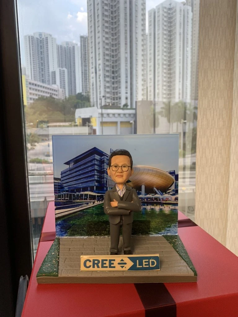 一位男性人像公仔站在石磚步道造型底座中央，雙手交叉、身穿深灰色西裝與條紋襯衫，配戴眼鏡，神情自信。背板為現代科技園區或創新中心風格的建築與水景照片。底座前方有藍白色牌匾，印有「CREE LED」字樣與箭頭圖示。整體呈現科技與照明產業氛圍的人像公仔展示。