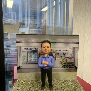 一款老闆主題的人像公仔，站在綠灰色砂石質感底座上，雙臂交叉、藍色長袖襯衫、黑色長褲與棕色鞋。底座前方名牌印有「Kenneth So / All the Best」與表情符號。背板是便利商店室內場景，包含飲料冷藏櫃、貨架與照明；整體置於臨窗環境，窗外可見高樓城市景觀。