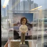 透明展示盒中的院長榮休主題人像公仔：短捲髮、戴圓框眼鏡的女性公仔，穿白色麻花針織上衣、米色長褲與白鞋，雙手交疊站立於方形底座。身後背板為教堂建築照片，屋頂有紅色十字架，周圍樹木花海；底座一角鋪有綠草與小白花。展示盒左前方立有中文感謝銘牌，提及榮休賀辭與「神召會聖光堂」。整體由手托起，背景是窗外城市高樓景色。