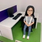 一款女性人像公仔坐在黑色辦公椅上，穿藍白條紋上衣、淺藍長褲與白色鞋，配戴眼鏡並微笑。旁邊為白色L形辦公桌與紫色桌屏，桌上擺有黑色螢幕、鍵盤、滑鼠與滑鼠墊。地台為草綠色質感，前方木質名牌刻有退休祝賀字樣。