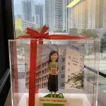 一款放於透明展示盒內並以紅色緞帶蝴蝶結包裝的退休公仔人像公仔。公仔留中長髮，穿淺綠外套、條紋上衣與深色長裙，雙手抱著一本橘色書本；腳邊有小型花束造型，站在綠色草地底座上。背景板為白色與紅色相間的中學校舍立面，右側可見「TAK OI SECONDARY SCHOOL」及中文校名。前方牌匾寫有「Enjoy Your 7-day Weekend!」。整體置於木紋桌面，窗外是高樓城市景觀。