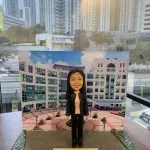 一款人像公仔作為退休公仔展示，公仔留長黑髮，穿黑色西裝外套與長褲、內搭淡粉上衣，站在灰色磚紋地台上；背景板印有白色與淺色系的多層學校/校園建築與中庭圓形花壇。底座前方牌匾寫有「Happy Retirement to Hidy 2024」。整體擺放在木紋桌面上，窗外可見高樓城市景觀。