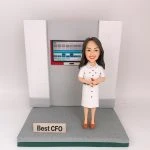 灰白色辦公背板前的CFO主題人像公仔：長深色髮女性公仔穿白色襯衫式連身裙，腳踩棕色尖頭鞋，雙手握紅色筆微笑站立。身後嵌入式螢幕顯示藍灰色表格與儀表板介面。公仔立於灰色紋理底座，前方放置金屬色小名牌寫有「Best CFO」。背景為純白。