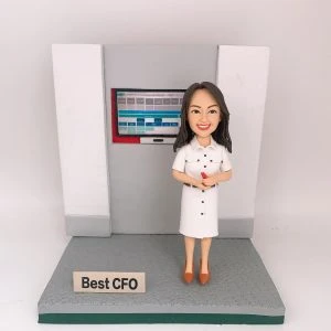 灰白色辦公背板前的CFO主題人像公仔：長深色髮女性公仔穿白色襯衫式連身裙，腳踩棕色尖頭鞋，雙手握紅色筆微笑站立。身後嵌入式螢幕顯示藍灰色表格與儀表板介面。公仔立於灰色紋理底座，前方放置金屬色小名牌寫有「Best CFO」。背景為純白。