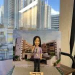 一款女性人像公仔站在灰色石磚質感的長方形底座上，身穿淡紫色上衣與深色長褲，胸前掛著識別證，右手比出V字手勢並微笑。背板為一幅校園建築照片，包含多棟相連的米白與棕色教學樓、窗戶與入口車道。底座前緣放有淺木色名牌，上有英文字樣「Happy Retirement」。整體擺設置於室內木桌上，窗外可見高樓大廈倒影與自然光。