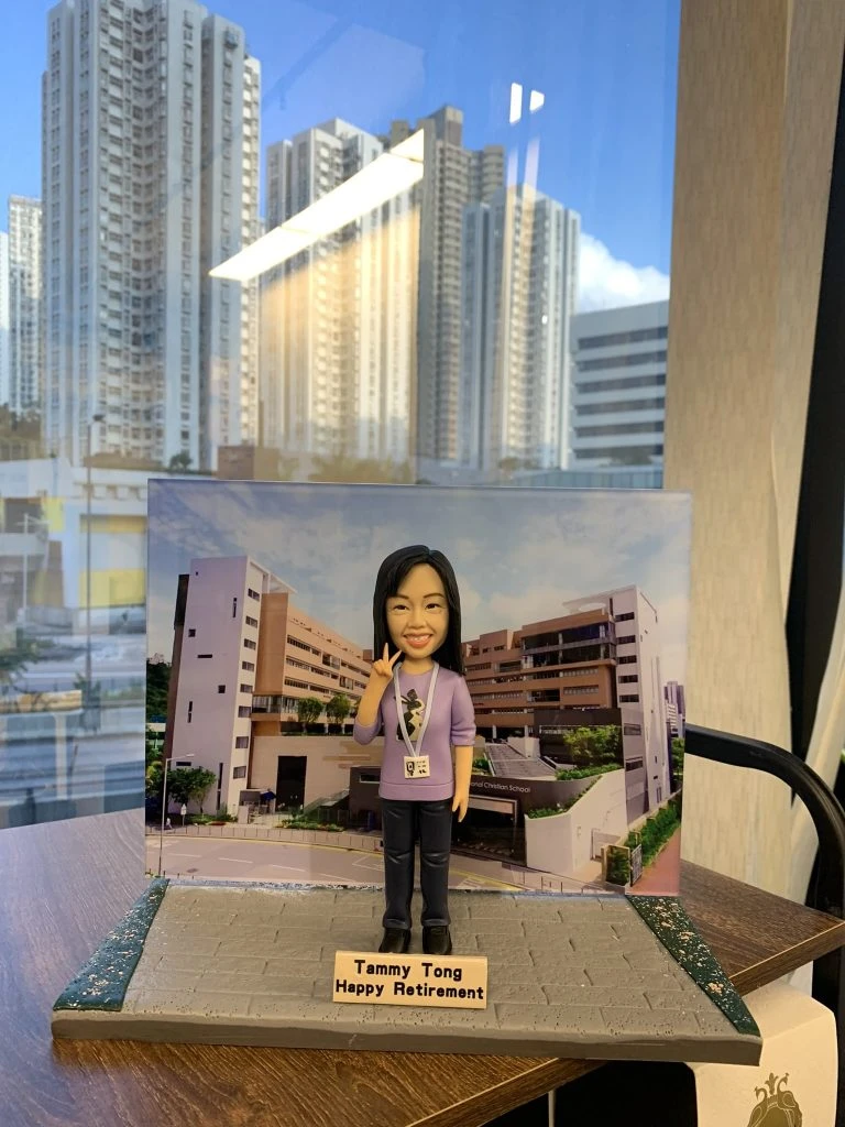 一款女性人像公仔站在灰色石磚質感的長方形底座上，身穿淡紫色上衣與深色長褲，胸前掛著識別證，右手比出V字手勢並微笑。背板為一幅校園建築照片，包含多棟相連的米白與棕色教學樓、窗戶與入口車道。底座前緣放有淺木色名牌，上有英文字樣「Happy Retirement」。整體擺設置於室內木桌上，窗外可見高樓大廈倒影與自然光。