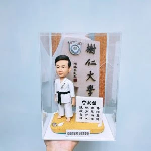透明展示盒中的人像公仔:短黑髮男性造型穿白色空手道道服、黑帶,站在沙色底座上,旁邊有小白石與海貝裝飾。右側立有白色立牌,上面以黑字書寫武道箴言。背板為校園入口石碑,上有「樹仁大學」字樣與校徽。展示盒前方貼有細長名條,寫有祝福語。背景為淺藍灰色牆面,由手掌托舉展示盒底座。