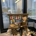 咖啡露台主題的人像公仔：男性穿棕色西裝外套、綠色針織背心與黑褲皮鞋，優雅坐在木椅旁的折疊木桌，桌上擺有紅酒瓶、酒杯、點心拼盤與向日葵裝飾；背板懸掛楓葉與足球隊徽旗幟，地台為彩色方塊磁磚。前方展示萬通保險（YF Life）名片，整體呈現同事升職慶賀、公司致贈禮的人像公仔。