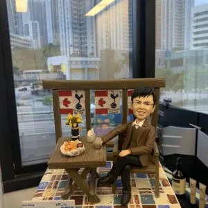 咖啡露台主題的人像公仔:男性穿棕色西裝外套、綠色針織背心與黑褲皮鞋,優雅坐在木椅旁的折疊木桌,桌上擺有紅酒瓶、酒杯、點心拼盤與向日葵裝飾;背板懸掛楓葉與足球隊徽旗幟,地台為彩色方塊磁磚。前方展示萬通保險(YF Life)名片,整體呈現同事升職慶賀、公司致贈禮的人像公仔。