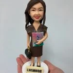 一款女性人像公仔被手掌托起展示。公仔留長深棕髮、微笑，身穿深棕色V領連身裙與黑色平底鞋；右臂抱著粉紅到藍綠漸層封面的冊子，左手提黑色手袋。站在白色圓形底座上，前方有淺米色名牌刻字「LWS創校功臣」。背景為素灰白色。