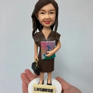 一款女性人像公仔被手掌托起展示。公仔留長深棕髮、微笑，身穿深棕色V領連身裙與黑色平底鞋；右臂抱著粉紅到藍綠漸層封面的冊子，左手提黑色手袋。站在白色圓形底座上，前方有淺米色名牌刻字「LWS創校功臣」。背景為素灰白色。