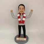 人像公仔立於圓形灰色底座上，雙手高舉呈慶祝姿勢；短髮、面帶笑容，穿灰色連帽上衣、黑色長褲與紅白運動鞋，頸上繫紅白相間條紋圍巾。底座前方金色銘牌印有英文字：「Steven Lee / 18 Years with AQUA!」。