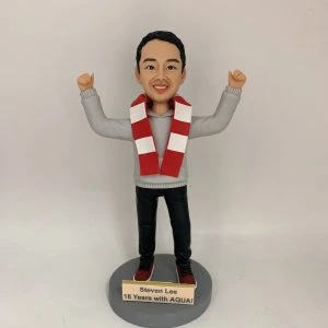 人像公仔立於圓形灰色底座上，雙手高舉呈慶祝姿勢；短髮、面帶笑容，穿灰色連帽上衣、黑色長褲與紅白運動鞋，頸上繫紅白相間條紋圍巾。底座前方金色銘牌印有英文字：「Steven Lee / 18 Years with AQUA!」。