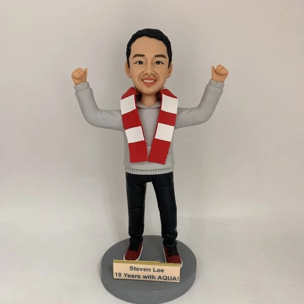 人像公仔立於圓形灰色底座上，雙手高舉呈慶祝姿勢；短髮、面帶笑容，穿灰色連帽上衣、黑色長褲與紅白運動鞋，頸上繫紅白相間條紋圍巾。底座前方金色銘牌印有英文字：「Steven Lee / 18 Years with AQUA!」。