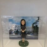 透明壓克力展示盒內，站在圓形草地底座上的女性人像公仔，黑色長髮，穿黑色印花上衣與深色過膝裙，雙手在身前交握。背板是一幢白色多層校舍與校門口的照片，門柱與牆面可見學校英文名稱「Secondary School」以及中文直書校名（畫面不全但清楚為中學校舍正門），天空藍天白雲，校門前有藍色圍欄與樹木。