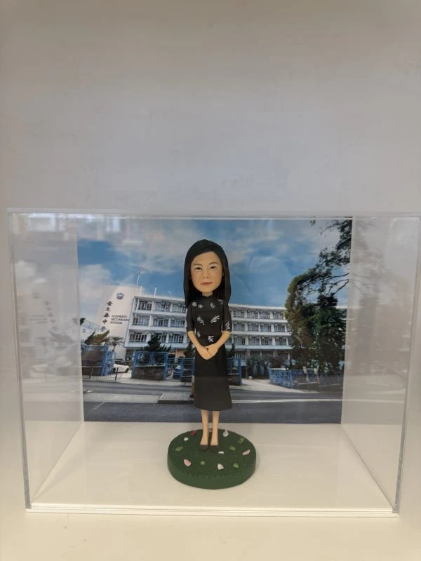透明壓克力展示盒內，站在圓形草地底座上的女性人像公仔，黑色長髮，穿黑色印花上衣與深色過膝裙，雙手在身前交握。背板是一幢白色多層校舍與校門口的照片，門柱與牆面可見學校英文名稱「Secondary School」以及中文直書校名（畫面不全但清楚為中學校舍正門），天空藍天白雲，校門前有藍色圍欄與樹木。