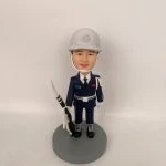 穿深藍制服與白色安全帽的男性人像公仔，戴白色頤帶與肩繩，胸前有彩色識別條與白色腰帶；右手持黑白配色禮槍，黑靴踏立於灰色圓形底座。背景為純白牆面。