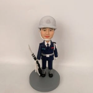 穿深藍制服與白色安全帽的男性人像公仔,戴白色頤帶與肩繩,胸前有彩色識別條與白色腰帶;右手持黑白配色禮槍,黑靴踏立於灰色圓形底座。背景為純白牆面。