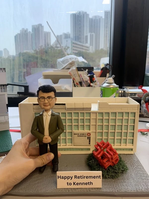 男性人像公仔穿白襯衫與綠色外套、黑長褲與黑鞋，雙手於身前交握，站在灰色情境底座上。背後是帶有「Housing Authority Headquarters」與「房屋委員會」標示的白色建築模型，右前方有紅色幾何雕塑與綠色草叢。底座前方立有白色名牌寫著「Happy Retirement to Kenneth」。畫面邊緣可見工作檯與城市窗景。