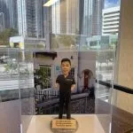 一款人像公仔置於透明展示盒中，公仔穿全黑牙科/醫護手術服，上衣左胸印有「Dr. Darren Yeo」字樣；背景是白色外牆、紅門與招牌的牙科診所場景。公仔站在圓形灰色底座上，前方金屬色牌寫著「Excellent Dentist • Amazing work done • Thank you Darren」。展示盒放在木紋桌面上，窗外可見高樓城市景觀。