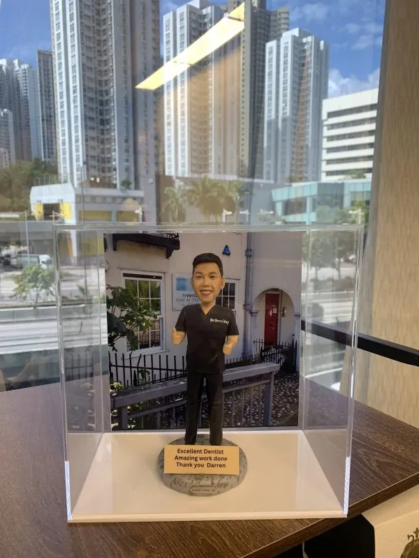 一款人像公仔置於透明展示盒中，公仔穿全黑牙科/醫護手術服，上衣左胸印有「Dr. Darren Yeo」字樣；背景是白色外牆、紅門與招牌的牙科診所場景。公仔站在圓形灰色底座上，前方金屬色牌寫著「Excellent Dentist • Amazing work done • Thank you Darren」。展示盒放在木紋桌面上，窗外可見高樓城市景觀。
