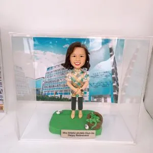 透明壓克力展示盒內，一尊女性人像公仔站在綠色草地質感底座上，短至中長髮、微笑，穿彩色幾何花紋上衣、深色及踝褲與米黃色鞋，雙手於身前交握。底座右側有一小簍綠植與白色小石裝飾。前方金色名牌刻有「Mrs CHAN LEUNG Chui-ha Happy Retirement」。背板是一張校園建築照片，左側為藍白相間的多層教學大樓，右側藍色牆面印有英文與中文校名「SHAU KEI WAN GOVERNMENT SECONDARY SCHOOL／筲箕灣官立中學」及學校盾形校徽，藍天白雲作背景。
